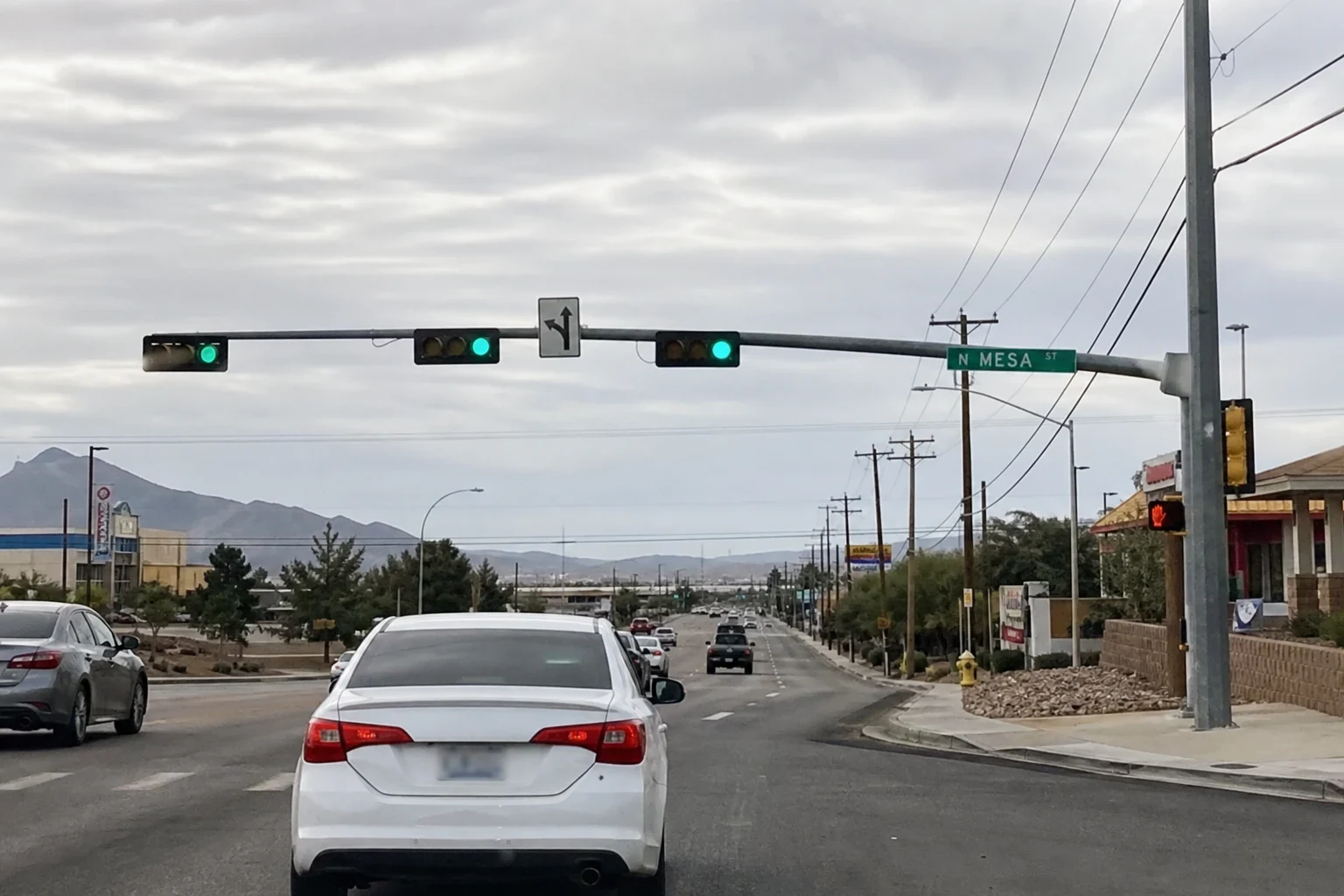 Overcast view of traffic on a wide arterial street at a green light, with a white sedan ahead, utility poles, low commercial buildings and mountains in the distance